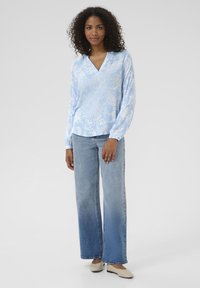 Light blue blouse with white paisley pattern, V-neck, and long sleeves, paired with wide-leg faded denim jeans and beige flat shoes.