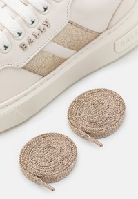 Białe skórzane sneakersy z teksturowanym złotym pasem oraz nazwą marki "BALLY"; obok buta znajdują się dwie spiralne złote sznurówki.