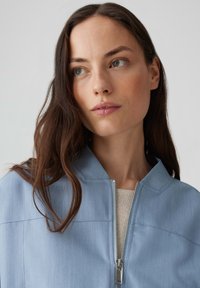 Veste zippée bleu clair avec un col montant et un tissu texturé, associée à un haut en tricot beige en dessous.