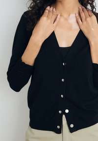 Svart cardigan i mjukt material med V-ringning, med knappstängning, långa ärmar och slät yta, som bärs över en matchande topp.