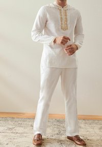 Uomo che indossa una tunica tradizionale bianca ricamata e pantaloni abbinati con mocassini in pelle marrone con nappe, in piedi su un tappeto decorato.