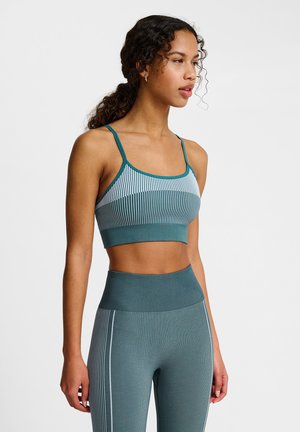 Kvinde med krøllet hår i teal- og lyseblåstribet sports-bh og højtaljede leggings, stående mod en ensfarvet baggrund.
