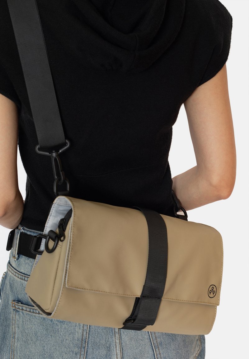 Béžová obdélníková kabelka přes rameno s černým popruhem, s magnetickým uzávěrem a detaily loga. Hladký materiál, minimalistický design.