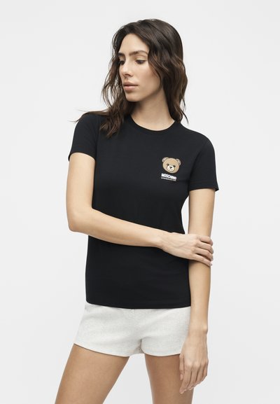Γυναίκα με σγουρά σκούρα μαλλιά που φοράει μαύρο T-shirt με μικρό σχέδιο αρκουδάκι και λευκά σορτς, ποζάροντας μπροστά σε λευκό φόντο.