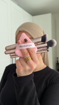 Set med fyra sminkborstar hållna i handen, med metallhandtag och mjuka strån. Följt av en rosa svampapplikator märkt "REVOLUTION".