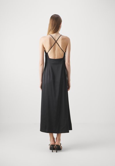 Abito lungo in satin nero con schiena profonda, sottili spalline incrociate e una texture liscia, abbinato a tacchi aperti.