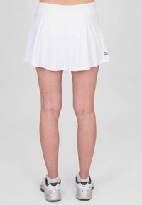 Falda atlética blanca con un diseño acampanado y una textura suave. Presenta una cinturilla ajustada y un detalle del logo cerca del dobladillo. Usada con zapatillas.