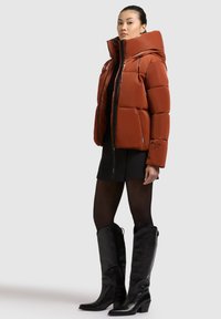 Kurtka puchowa w ciemnym pomarańczowym kolorze z wysokim kołnierzem, zamkiem błyskawicznym z przodu oraz pikowanym wzorem; zestawiona z czarną spódnicą i czarnymi botkami do kolan.