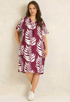 Kvinde iført en løs, knælængde, mørkerød kjole med store hvide bladmotiver og hvide sneakers, stående mod en ensfarvet væg.