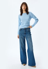 Hellblauer, weicher Strickpullover mit Rundhalsausschnitt. Kombiniert mit weit geschnittenen, verblassten blauen Jeans aus Denim, die über eine Taschen und einen Knopfverschluss verfügen.