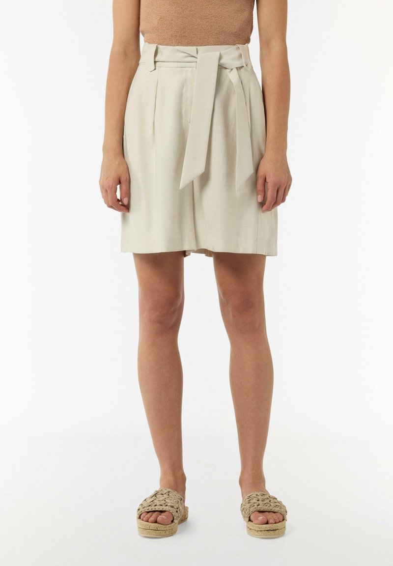 Light beige high-waisted shorts with a front tie, pleats, and side pockets. Paired with woven beige slip-on sandals. Smooth fabric texture.