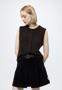 Bezruký hnedý pletený top s prednými gombíkmi, v kombinácii s tmavou kordurovou sukňou s záhybmi. Čierny opasok dodáva kontrast.