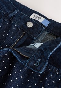 Laiade säärega kõrge vöökohaga teksad tumedas sinises denimis, mis on kaunistatud hõbedaste naeltega, ees tõmblukk ja metallist nööpkinnitus. Sisemine silt.