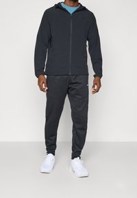Homme portant un sweat à capuche noir zippé, un pantalon de jogging noir et des chaussures de sport blanches, debout devant un fond clair uni.
