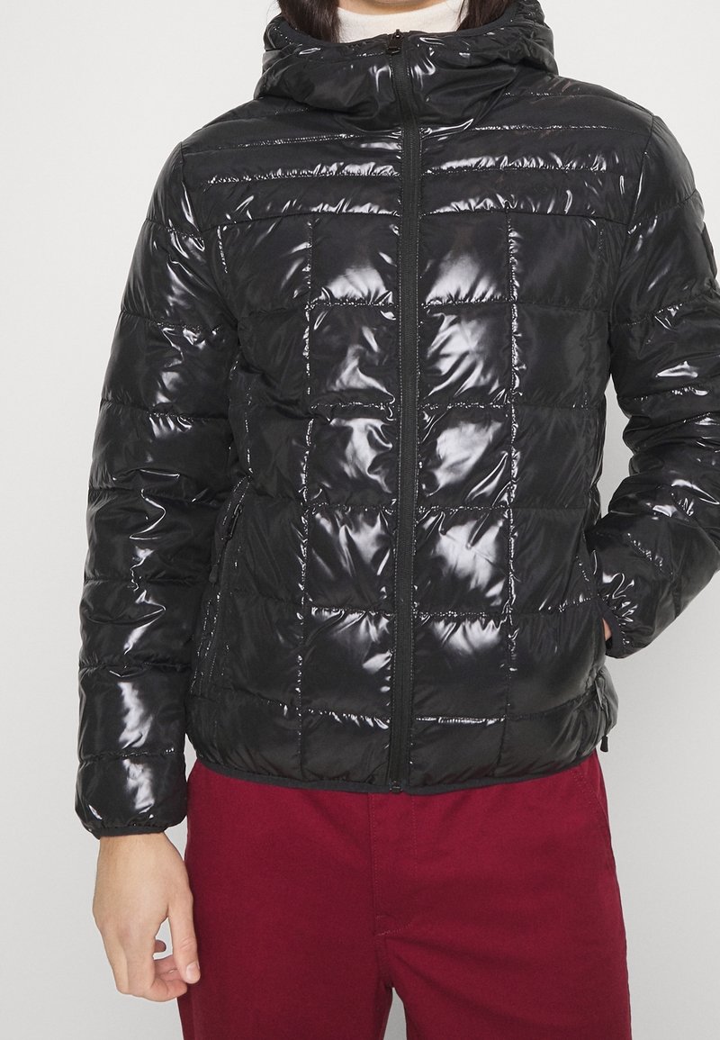 Veste matelassée noire brillante avec capuche zippée, portée par une personne avec une main dans la poche et un pantalon rouge visible de la taille jusqu'en bas.