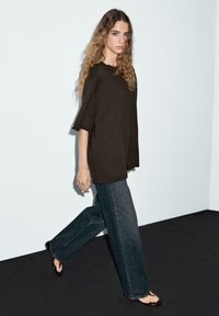 Maglia in maglia di colore marrone scuro oversize con maniche larghe, abbinata a jeans in denim scuro larghi. Il modello indossa ciabatte nere. Sfondo minimalista.