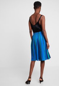 Czarny top bez rękawów z głębokim dekoltem na plecach w kształcie litery V, w połączeniu z plisowaną spódnicą w pionowe niebieskie i zielone paski. Czarne sandały na obcasie.