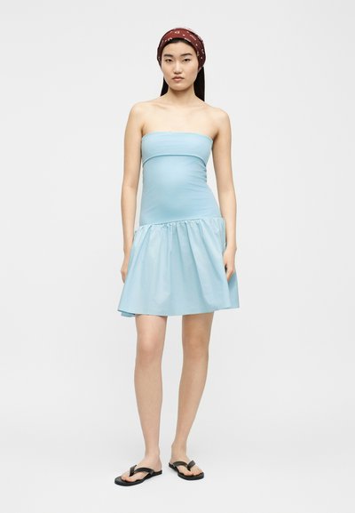 Νεαρή γυναίκα που φοράει ανοιχτό μπλε φόρεμα χωρίς τιράντες, μαύρα σαγιονάρες και κόκκινη κορδέλα με σχέδια, στέκεται μπροστά σε απλό φόντο.