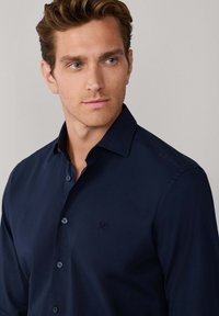 Camisa azul marino con botones y cuello puntiagudo, que presenta un sutil logotipo bordado en el pecho y una textura de tela suave.