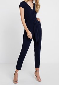Mono azul marino con mangas cortas, escote en V, cintura ceñida por un cinturón, patas ajustadas y una textura suave. Lleva tacones plateados con tiras.
