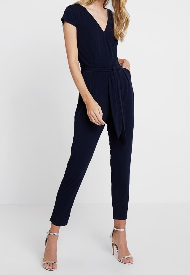 Mono azul marino con mangas cortas, escote en V, cintura ceñida por un cinturón, patas ajustadas y una textura suave. Lleva tacones plateados con tiras.