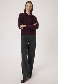 Donna che indossa un cardigan bordeaux con bottoni, jeans neri a gamba larga e décolleté neri a punta, in piedi con le braccia incrociate su uno sfondo neutro.
