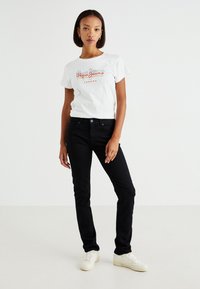 Camiseta blanca de manga corta con texto en logo rojo y negro, combinada con pantalones negros ajustados y zapatillas blancas. Atuendo simple y casual.