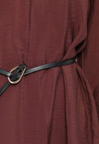 Μαύρη δερμάτινη ζώνη με χρυσό μεταλλικό κυκλικό διακοσμητικό, φορεμένη πάνω από ένα μπορντό υφασμάτινο ρούχο με υφή, που αναδεικνύει λεπτομέρειες με δούλεμα.