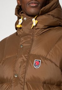 Brown puffer jacket featuring a high collar, snap buttons, and a yellow drawstring. Includes a logo patch on the chest with a red fox.