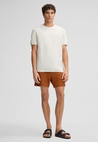 Hellbeige T-Shirt mit kurzen Ärmeln, braune strukturierte Shorts und schwarze Sandalen. Einfacher, lässiger Schnitt mit Rundhalsausschnitt.
