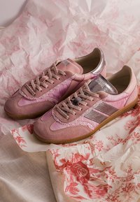 Paire de baskets roses en daim et tissu avec lacets beige, accents métalliques rose et semelles en gomme reposant sur un papier de soie à motif floral.