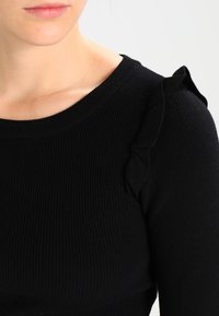 Haut en tricot noir à côtes avec un col rond et des accents froncés aux épaules. Texture lisse avec une coupe ajustée, épaisseur moyenne.