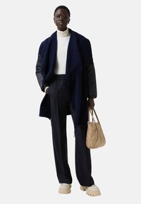 Námořnický pletený šál s třásněmi, přehnutý přes bílé rolákové triko. Slim fit námořnické kalhoty a béžová prošívaná kabelka. Silné krémové tenisky.