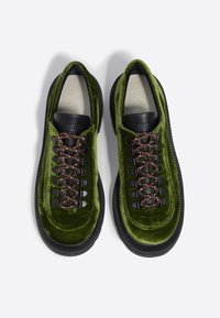 Zapatillas de terciopelo verde con detalles en cuero negro y cordones marrones. Presentan una superficie texturizada y una suela gruesa de goma negra.