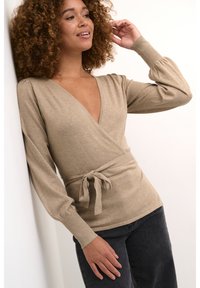 Top envolvente beige con un profundo escote en V, mangas largas abullonadas y un detalle de cintura atada. Hecho de una tela suave con una textura lisa, combinado con jeans oscuros.