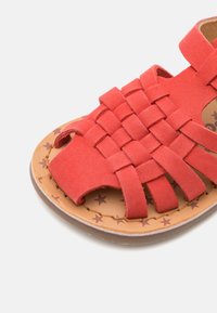 Røde semskede sandaler med et vævet overdesign, åben tå og en brun indersål med stjernemønstre. Akkompagneret af en flad gummisål.