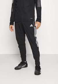 Person med svarta Adidas träningsbyxor med vita ränder och logotyp, i kombination med svart- och vitfärgade sportskor, står på ett vitt golv.