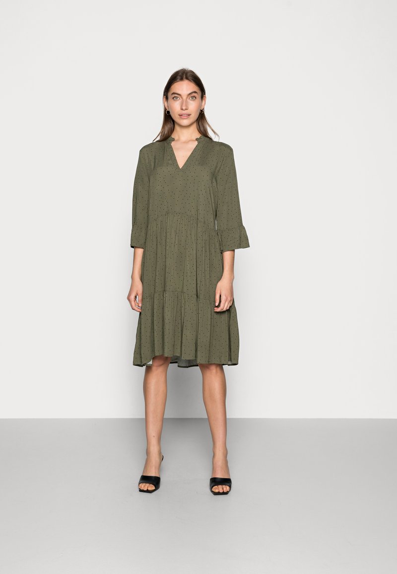 Vestido verde-oliva com mangas três quartos, decote em V e saia em camadas. Apresenta um padrão de pontos subtil e tecido fluido. Usado com sapatos de salto pretos.