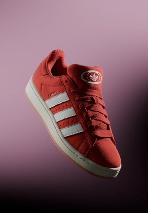 Basket rouge Adidas Campus avec des bandes blanches et une semelle en gomme, inclinée sur un fond dégradé violet et noir.
