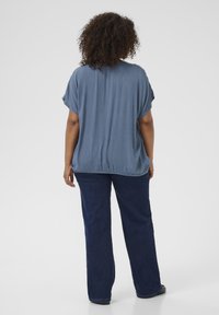 Frau mit lockigem Haar, die ein lockeres blaues Kurzarmoberteil und eine dunkelblaue Jeans trägt, steht mit dem Rücken zu einem schlichtem Hintergrund.