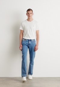 Tricou alb din bumbac, blugi de denim albaștri și adidași albi. Modelul stă în fața unui fundal simplu, având o croială lejeră și un design simplu.