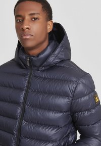 Giacca puffer navy con un design trapuntato e chiusura zip frontale. Presenta un cappuccio e una piccola toppa con il marchio sulla manica. Texture liscia e lucida.