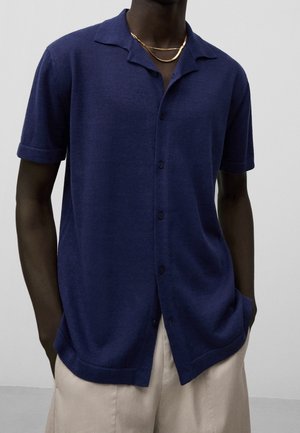 Mężczyzna w granatowej koszuli na krótki rękaw zapinanej na guziki i beżowych spodniach, ozdobiony dwoma złotymi łańcuszkami.