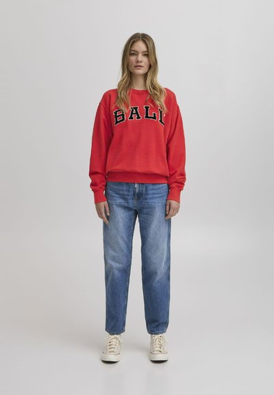 Rød hettegenser med "BALI" trykket i svart, kombinert med lyseblå straight-leg jeans og hvite joggesko. Glatt stoff med ribbet mansjetter.
