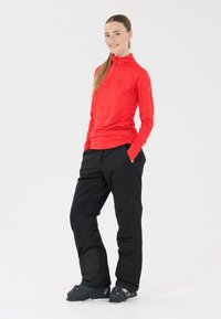 Rode lange mouw top met een kwart-rits en zwart gevoerde broek. Donkere laarzen met hardwareaccenten. Effen witte achtergrond.