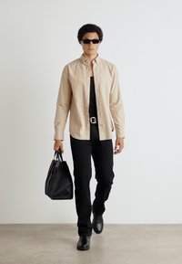 Camicia beige a maniche lunghe con bottoni sopra un top nero, abbinata a pantaloni neri attillati e scarpe. Tiene in mano una borsa nera con texture.