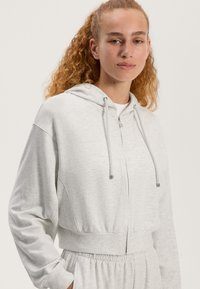 Zastrihnutá šedá mikina s kapucňou vyrobená z mäkkej tkaniny, so zipsom, šnúrkami a elastickým pásom. Obsahuje raglanové rukávy a textúrovaný povrch.