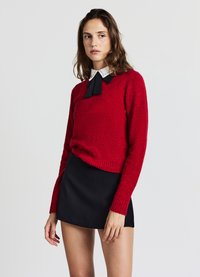 Roter Strickpullover mit langen Ärmeln und gerippten Bündchen, ausgestattet mit einer schwarzen Fliege und weißem Kragen, kombiniert mit einem schwarzen Minirock.