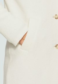 Manteau en laine blanche texturée avec une poche latérale et un grand bouton crémeux. Le tissu semble doux et chaud.