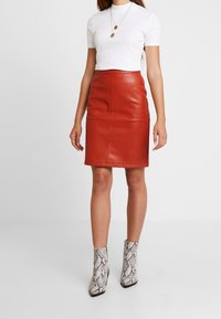 Červená kožená tužková sukně se švy, spárovaná s upnutým bílým ribovým topem a kotníkovými botami s hadím vzorem.